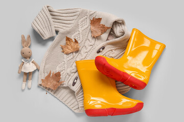 Yellow gumboots with sweater, toy bunny and autumn leaves on grey background