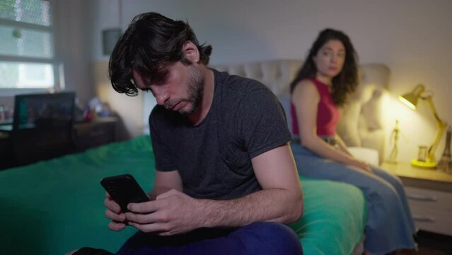 Disconnected couple: Boyfriend looking at phone ignoring girlfriend by in bedroom. Uncommunicative man absorbed by social media online while woman wants attention