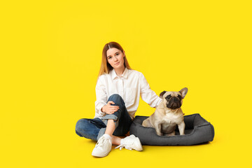 Beautiful young woman and cute pug dog sitting on pet bed against yellow background