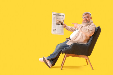 Mature man with pointing at newspaper and sitting on armchair against yellow background