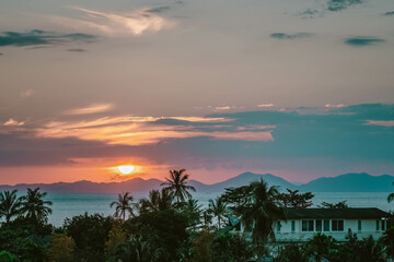 Fototapeta premium Scenic rose golden glowing Sunset. Half Sun hid in clouds above mountains. Close up view on palms, sea and mountains at horizon line. Ao Nang, Thailand