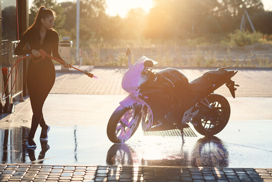 Young woman washes a motorcycle at self service car wash with high pressure water.