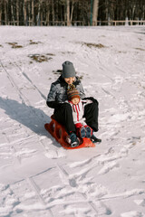 Fototapeta na wymiar Smiling mother with a small child sledding down a snow-covered hill