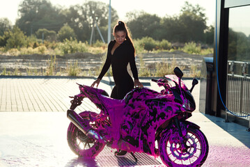 Pretty girl in black seductive suit stands near motorcycle at self-service car wash at sunrise.