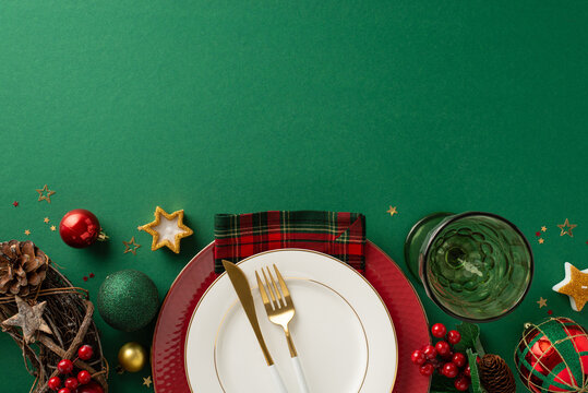 Christmas dinner table concept. Top-view image of plates, gilded flatware, plaid napkin, wine goblet, ornaments, rustic wreath, star candle, glistening confetti, mistletoe on green with room for text