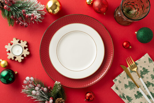 Elegant New Year's family dinner table setting with top-down view. Charger plate, gold cutlery, wine glass, baubles, candle, and festive decor on a red backdrop. Ample space for text or advertising