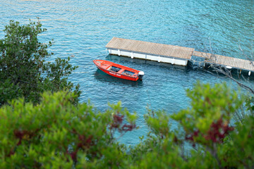 View to calm sea bay from above through some coniferous trees, red rescue boat docked near small wooden pier