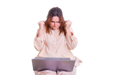 An adult woman is angry while sitting on the bed with a laptop, isolated on a white background. Angry woman raised her hands clenched into fists while working online from home