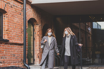 Naklejka premium Two young stylish women in medical masks talking and laughing while walking the street in town. Friends strolling outdoor in city