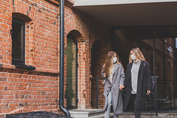Two young stylish women in medical masks talking and laughing while walking the street in town. Friends strolling outdoor in city
