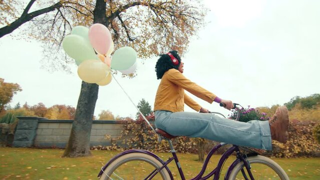 Chirpy female on a ladies bike with balloons attached to the trunk moving slowly to the open gate along wet path, listening to the music in headphones. High quality 4k footage