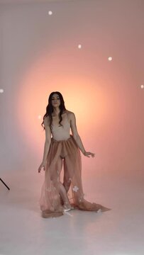 A young woman, a ballerina girl dancing, poses in a photo studio at a photo shoot, gently moving her arms and fingers on a pink background, slowmo, medium. Lady brown-haired woman with long hair