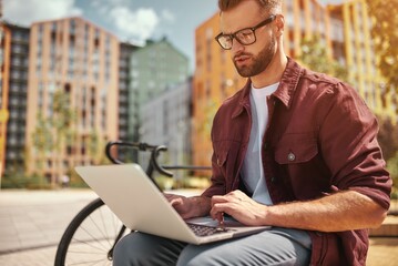 Modern technologies. Portrait of young man with stubble in casual clothes and eyeglasses working on laptop while sitting on the bench near his bicycle