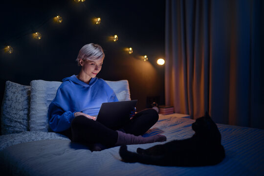 Young woman with her cat using laptop computer in bed in her cozy bedroom at night.