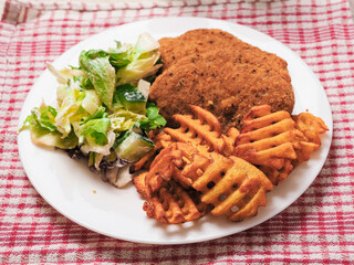 Two southern fried chicken fillets, green fresh salad and fancy cut potato lattice served on a white plate. Poultry meat dinner. Food served on a table.