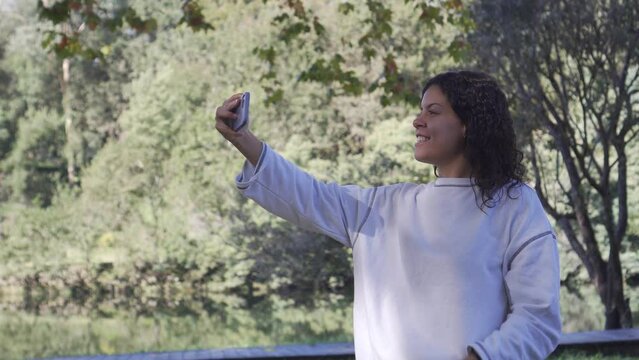 Young Caucasian brunette girl in white sweatshirt taking a smiling selfie with her cell phone in a natural setting