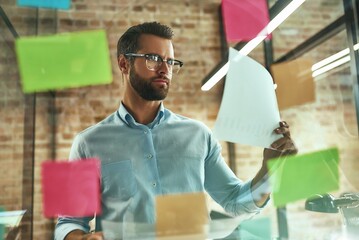 Creativity. Young and handsome bearded man in eyeglasses planning working process and using colorful stickers while standing in front of glass wall