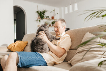 A middle-aged man sits and cuddles with a gray fluffy cat on the sofa in the living room and...