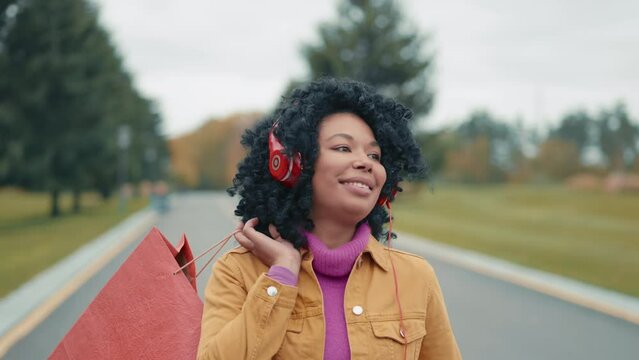 Happy dark-haired curly black woman wearing red headphones walking alone along roadway, holding red paper bag over the shoulder, admiring beauty of nature. High quality 4k footage