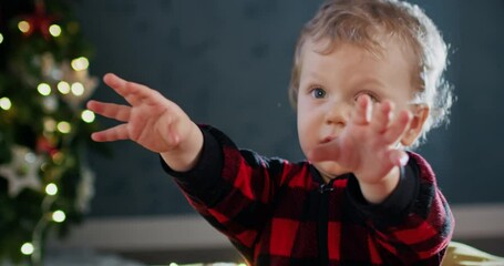 Toddler boy raises hands reaching to favourite Christmas present against tree with garland. Kid wants to open presents during New Year celebration