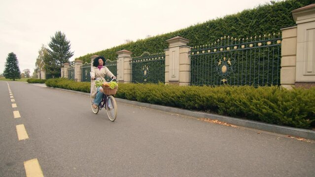 Romantic young lady riding a bicycle with basket full of products, looking with interest at luxurious metal fence with dense row of evergreen trees. High quality 4k footage