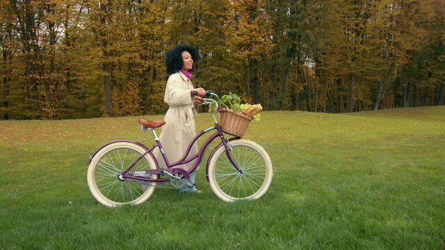 Nice African female strolling with ladies bike with attached basket on green lawn with autumn trees, observing picturesque landscape, breathing fresh air. High quality 4k footage