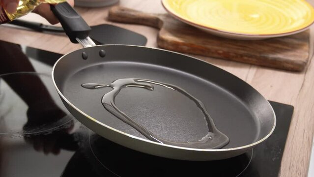 Pouring oil into frying pan. A close-up view captures the moment oil is being poured from a clear bottle into a black frying pan, with a wooden surface and a yellow plate in the background.