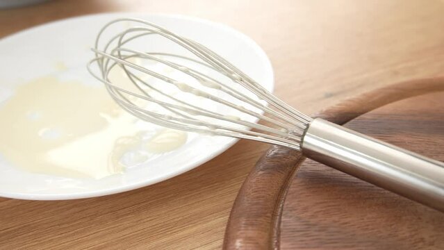 Whisk on wooden surface. A metal whisk rests on a wooden cutting board next to a white plate with remnants of food batter, illustrating a cooking or preparation scene.