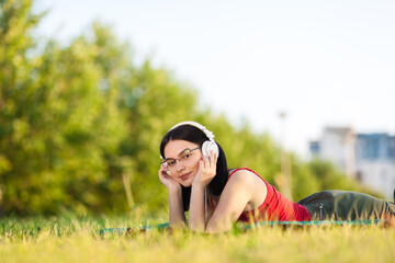 Beutiful dark hair girl listening to music with a headphones from her smartphone. Lying on a meadow in the park. Looking at camera.