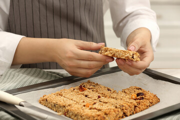 Woman taking granola bar from baking tray at table in kitchen, closeup