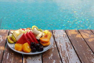 Plate with fresh fruits on wooden deck near outdoor swimming pool, space for text. Luxury resort