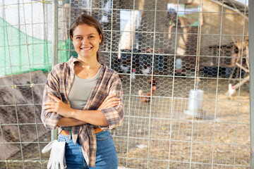Fototapeta premium Girl worker of farm standing after work. Highly specialized agricultural production, mini poultry farm. On hot afternoon, owner of livestock farm stands near corral mesh fence for livestock and rests
