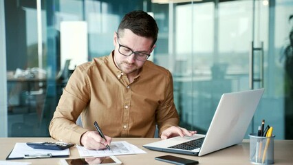 Busy financier or accountant making financial calculations using typing on a laptop computer, taking notes sitting at workplace. Confident businessman working fills out documents in business office - Powered by Adobe