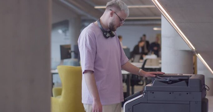 A modern, blond-haired man efficiently operates a high-tech paper printer in a large, contemporary office, showcasing advanced office technology and workflow management