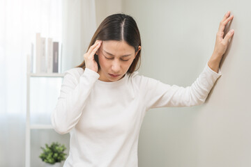 Dizzy asian young woman, girl headache or migraine pain. suffering from vertigo, difficulty standing up leaning on wall at home, holding head with hand, health problem of brain, inner ear not balance. - obrazy, fototapety, plakaty