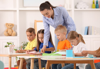 Nursery teacher with group of cute little children drawing and cutting paper at desks in kindergarten. Playtime activities