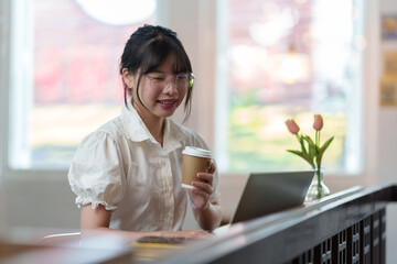 Asian female office worker Hold a hot cup of coffee, relax, and chat online. Internet via laptop computer While happily sitting at the counter in the library, office, or coffee shop.