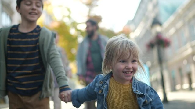 Happy little girl holding hands with her brother and walking in the city with parents behind them