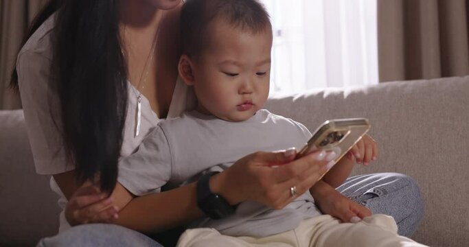 Mom and child on sofa looking to smartphone. Cinematic Chinese, Japanese, Korean, Asian. Baby looks enthusiastically at phone while sitting on his mother's lap. Leisure in happy family, emotional bond