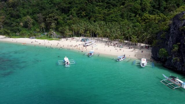 Seven Commandos palm beach in El nido with tourists on white sand swimming in blue water and Outrigger tour boats moving in foreground. Aerial