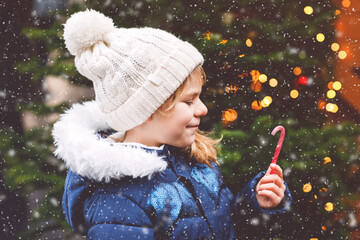 Little cute preschool girl with candy cane from a sweets stand on Christmas market. Happy child on traditional family market in Germany. Preschooler in colorful winter clothes during snowfall - Powered by Adobe