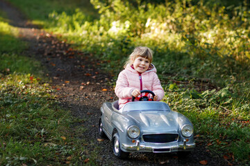 Little preschool girl driving big vintage toy car. Happy child having fun with playing outdoors. Active preschooler child enjoying warm autumn day in forest. Smiling stunning kid playing