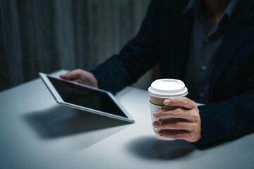 Businessman working in corporate workplace having reading data document on ipad screen while holding drinking coffee cup sitting in low light dark indoor office as young adult executive