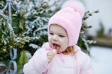 Portrait of little toddler girl walking outdoors in winter. Cute toddler eating sweet lollypop candy. Child having fun on cold snow day. Wearing warm baby pink clothes and hat with bobbles.