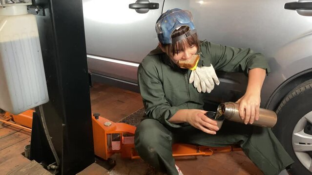 Young adult woman in coveralls at a car service station takes a well-deserved work break, savoring her lunch with a delicious sandwich and a hot beverage, providing a refreshing pause in her busy day