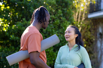 Happy diverse couple holding yoga mats talking in sunny garden