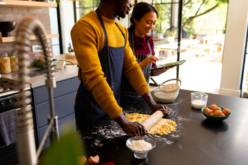 Happy diverse couple baking preparing dough and using tablet in sunny kitchen