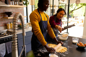 Happy diverse couple baking preparing dough and using tablet in sunny kitchen