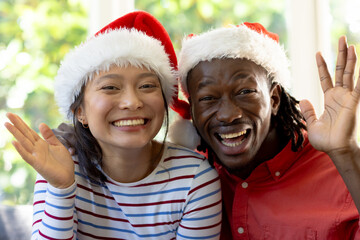 Happy diverse couple in christmas hats having video call waving in sunny living room - obrazy, fototapety, plakaty