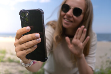 Young woman in sunglasses with shell bracelets holding smartphone. Influencer Using mobile phone for video call selfie waving on the beach seacoast. Holiday vacation advertisement concept. Social